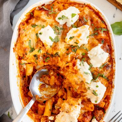 Baked Ziti with Sausage is baked in a white oval baking dish. There is a serving spoon in the dish that has scooped out a couple of servings of the baked ziti already.