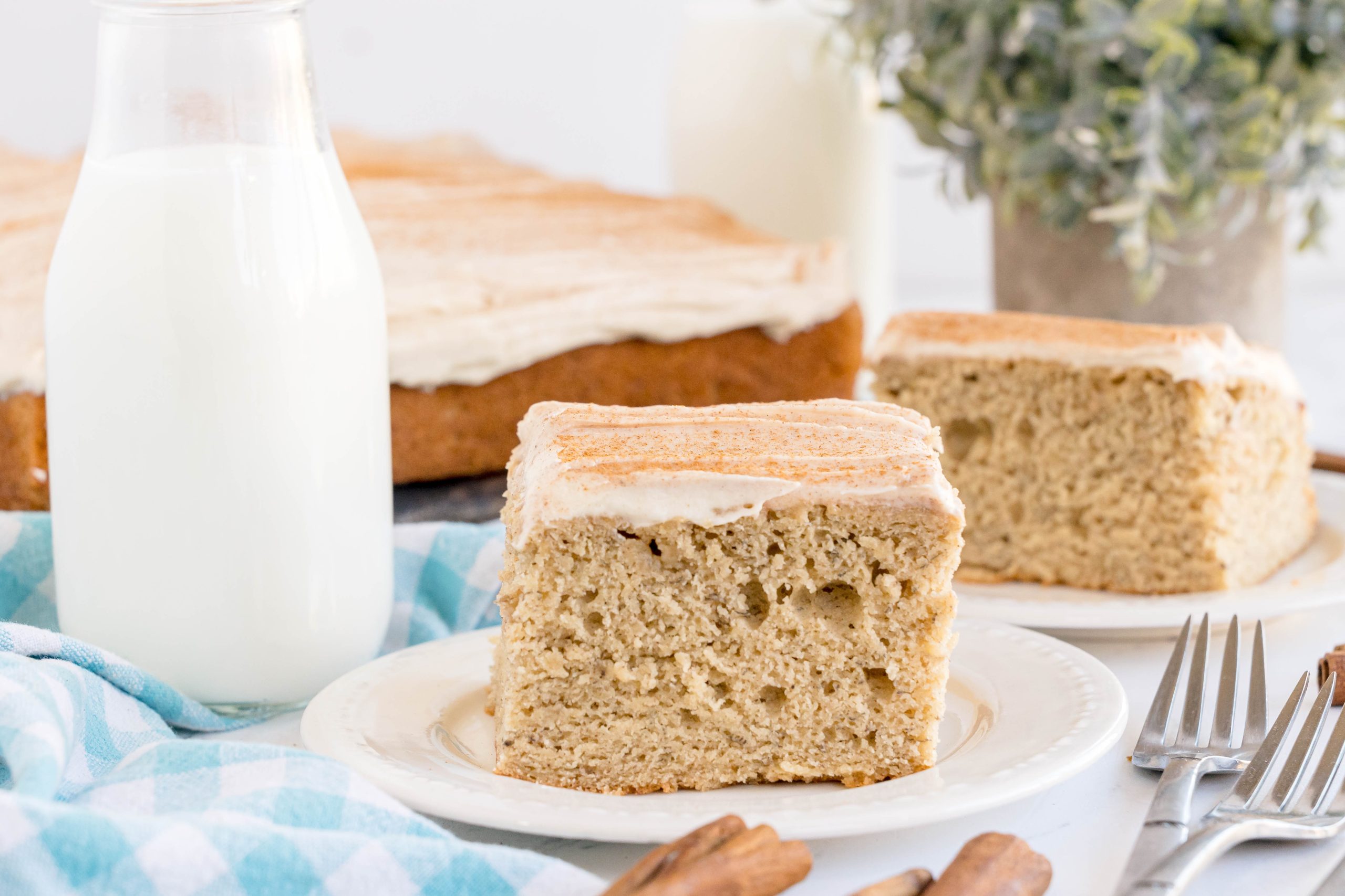 A piece of Banana Cake with Brown Butter Cream Cheese Frosting has been cut and is served on a white plate. There is more banana cake and a pitcher of milk in the background.