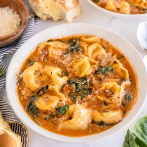 A bowl of Creamy Sausage Tortellini Soup is being served in a white round bowl. There is a buttered dinner roll and extra Parmesan cheese in a bowl near the soup.