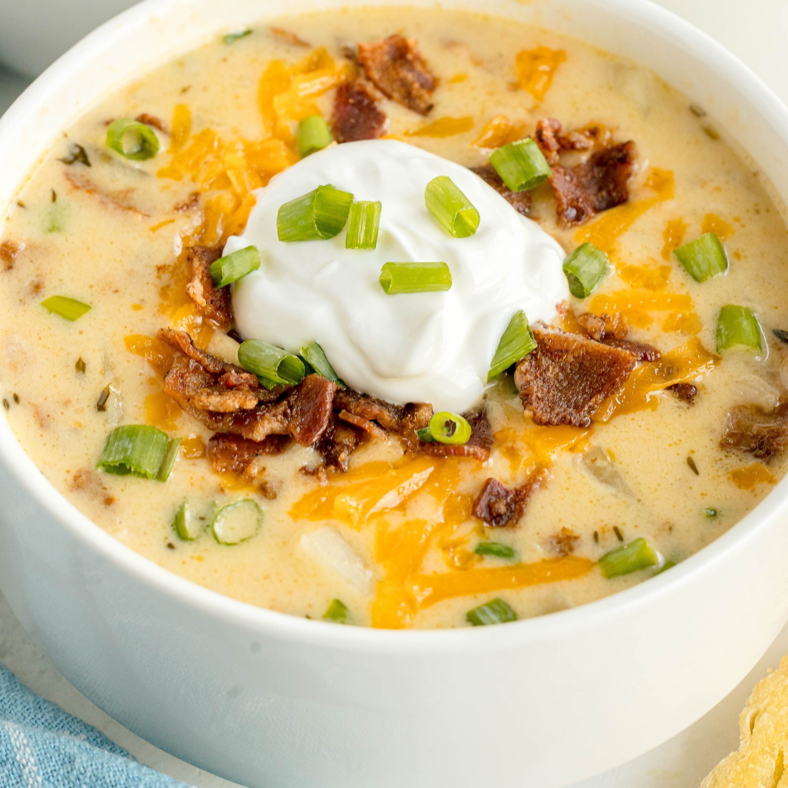A white bowl is full of Crock Pot Loaded Baked Potato Soup. On top of the soup is a dollop of sour cream, crumbled bacon, shredded cheddar cheese, and sliced scallions.